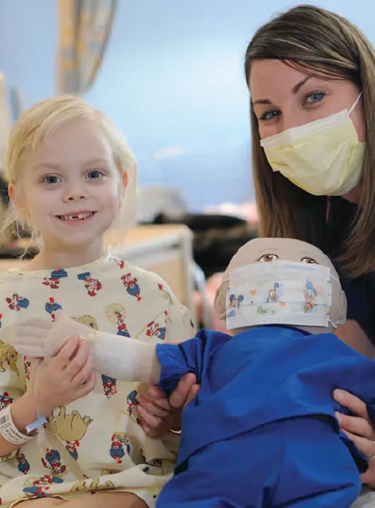 A young girl and a nurse smile as they both hold a doll wearing a surgical mask