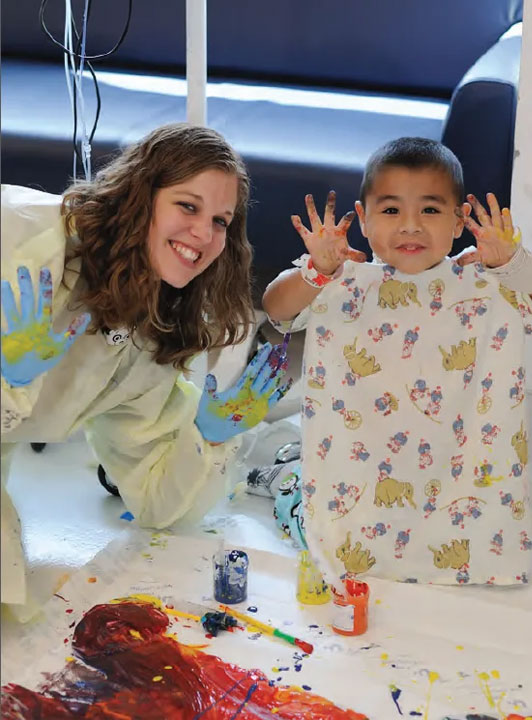 A nurse and a young boy patient both smile and hold up their hands to show themselves covered in paint.