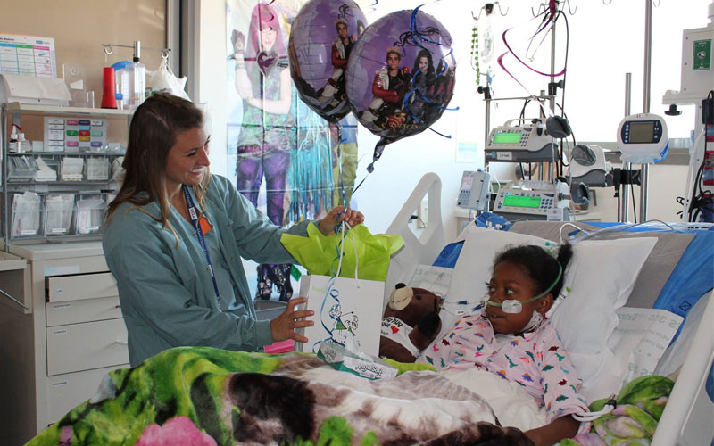 A young girl patient lying on a hospital bed and a nurse handing her a gift and balloons