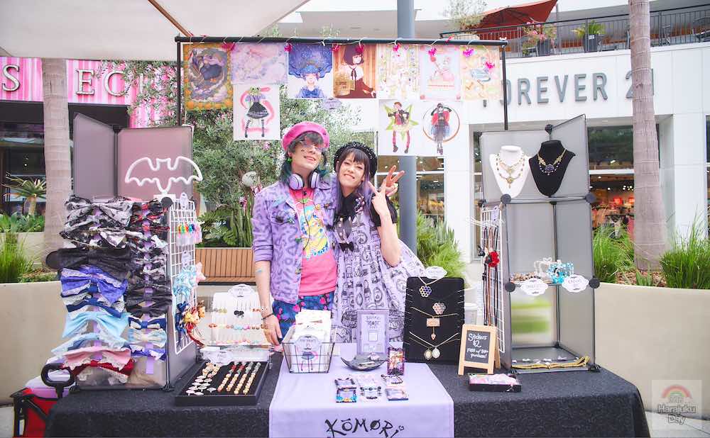 Two figures dressed in lavendar, patterned clothing standing behind their merchandise booth selling cute accessories and drawings.