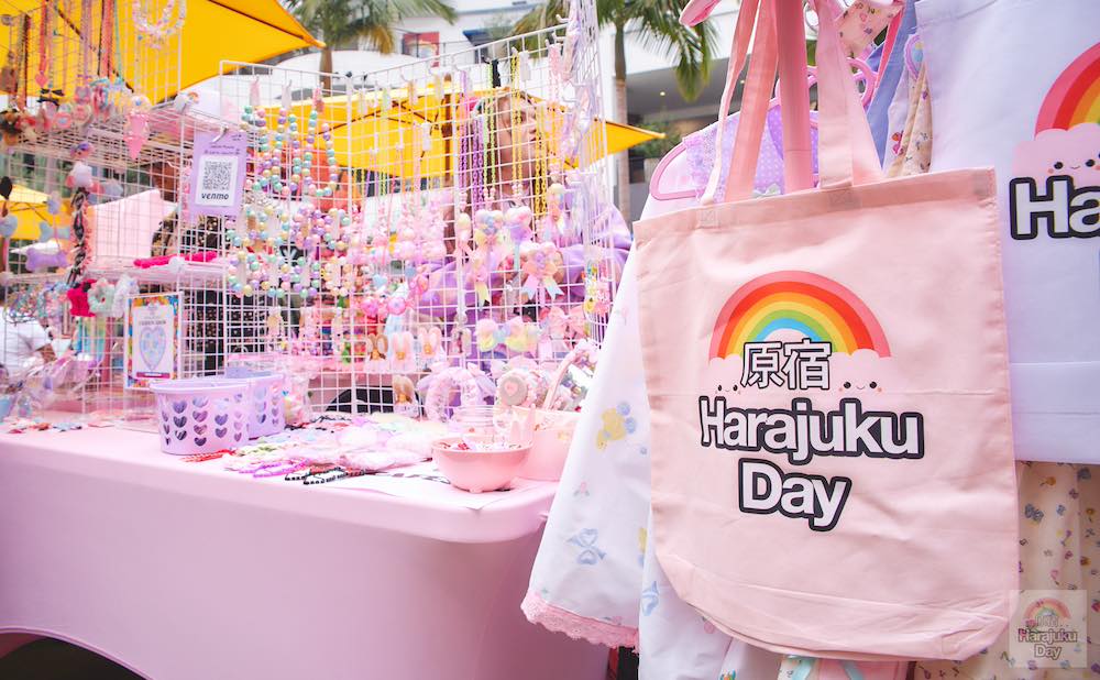 Pastel pink tote bag that says Harajuku Day hanging in front of a shop booth selling cute accessories.
