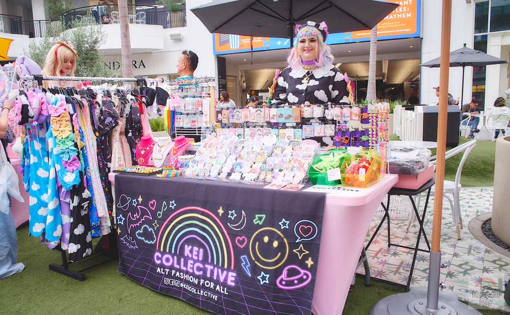 A shop owner in black clothing pattered with white clouds and pastel rainbow hair stands behind their cute fashion and accessories shop table