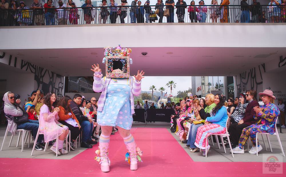 A fashion model with an old-school computer monitor for the head decorated with cute plushies wears a pale blue dress and poses for Kawaii Fashion Show