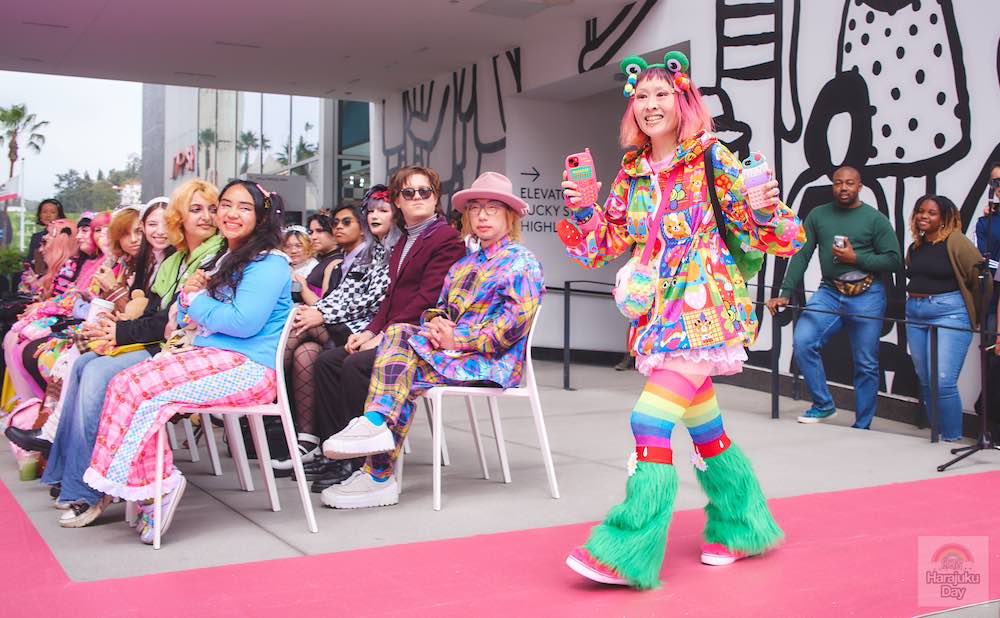 A fashion model in vividly, bright patterned clothing with frog headband and two colorful cellphones in hand walk during Kawaii Fashion Show