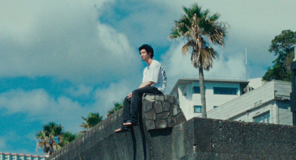 Still from movie "Super Happy Forever". Image shows a Japanese man sitting at the edge of a stone wall with blue skies and a palm tree in the back.