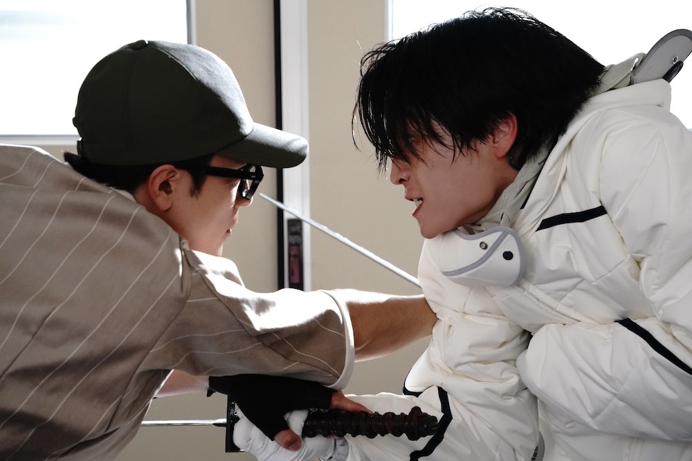 Film still from "Under Ninja." Two young men, one in a white down coat and the other in gray worker uniform with a black hat grapple with each other in a close combat fight.