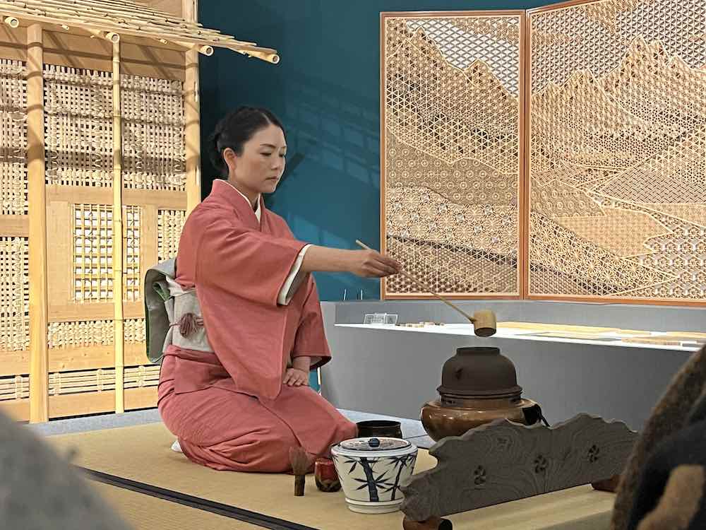 A woman in a salmon colored kimono sits in front of a Japanese tea house to perform a tea ceremony.