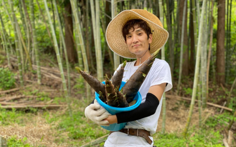 A man wearing a straw hat, holding a bucket filled with bamboo shoots (takenoko) in a bamboo forest.