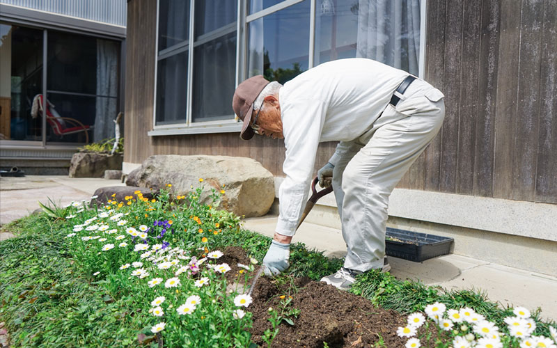 An elderly man gardening in his yard outside a wooden house.