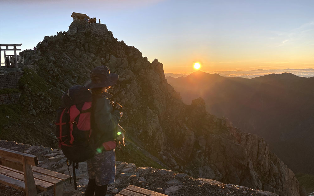 A hiker with a backpack gazing at a stunning mountain sunrise on a rocky peak.