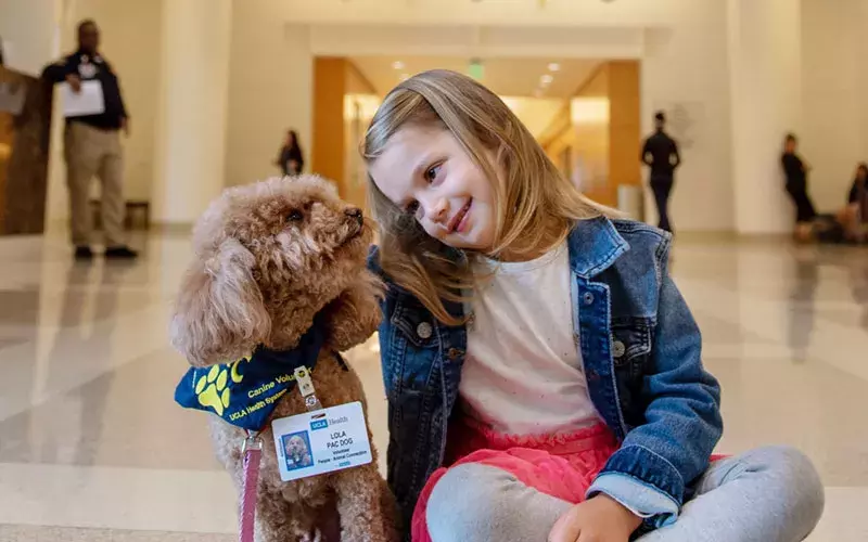 A young girl sits on the floor and smiles at the dog next to her.