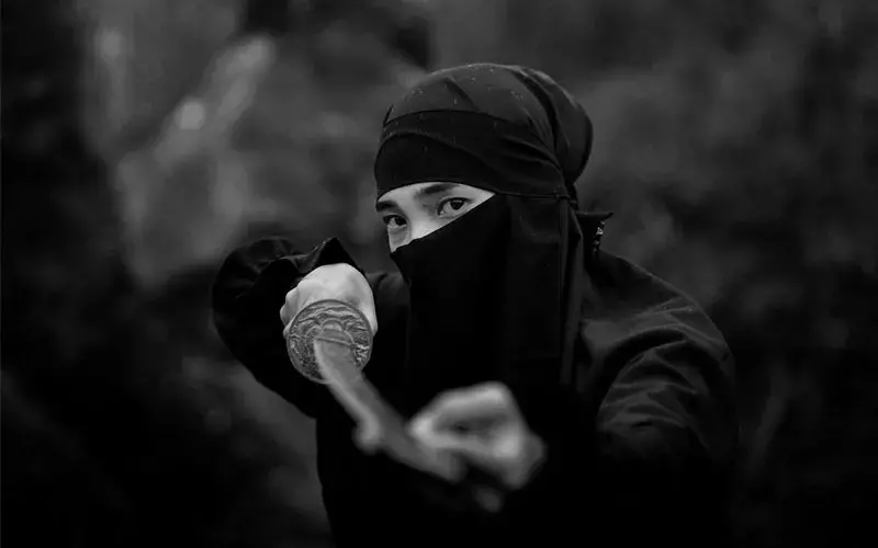 A man in ninja outfit holding a sword, black and white.